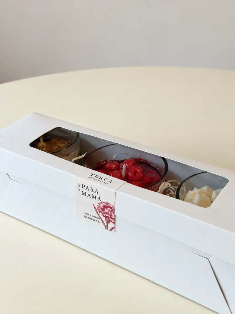 La caja de regalo ANTOJO cerrada sobre una mesa, con las tres velas visibles por la ventana y la faja PARA MAMÁ